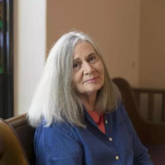 Marilynne Robinson sits on a pew and turns to face the camera, while light from a stained glass window shines on her. She has grey shoulder length straight hair, fair skin, and wears a blue button-down shirt with coral undershirt.
